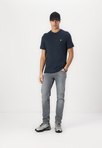 Mørkeblå bomull t-skjorte med liten gul logo, lys grå jeans og grå treningssko. Modell med svart caps.