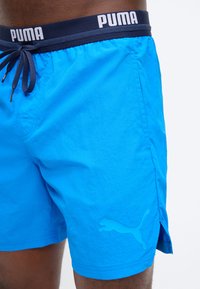 Άνδρας που φοράει φωτεινά μπλε μαγιό Puma με μπλε σκούρο ζωνάρι στη μέση και κορδόνι, με το λογότυπο Puma ορατό στο πόδι και στη μέση.