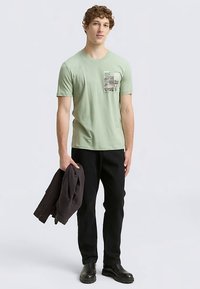 Hele-roheline puuvillane T-särk rinnal graafikaga; kombineeritud mustade pükste ja vasakus käes hoitava tumeda jakiga. Mustad saapad.