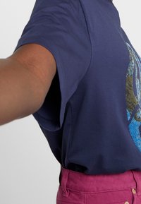 Person iført en navy blå kortærmet skjorte med et blåt og guld design, og højhalset fuchsia bukser, arm strakt fremad.