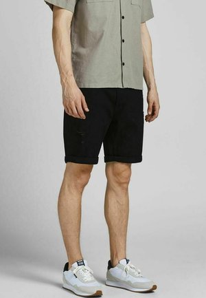 Mann trägt hochgekrempelte schwarze Shorts, hellgraues kurzärmeliges Hemd mit Knöpfen und weiße Turnschuhe, stehend vor einfarbigem Hintergrund.