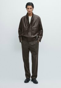 Giacca di pelle marrone con colletto alto e due tasche frontali, abbinata a pantaloni marrone a righe e scarpe nere. Design semplice e texturizzato.