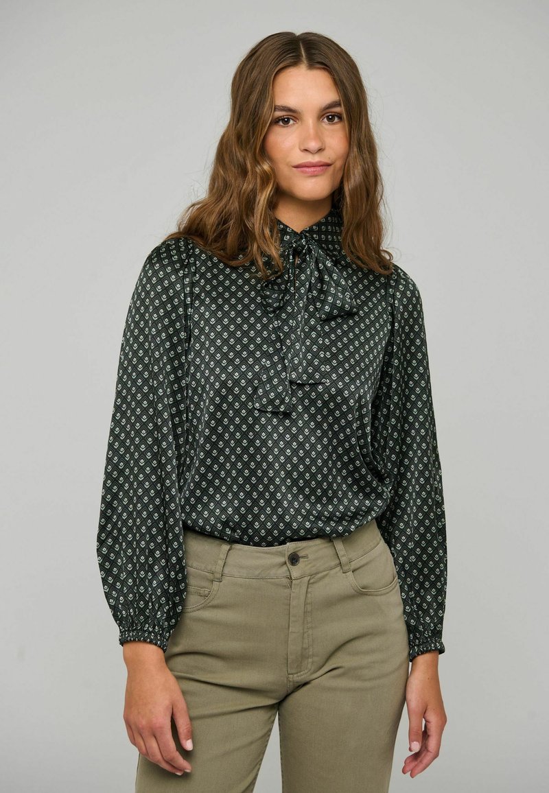 Jeune femme aux longs cheveux ondulés porte un chemisier noir à motifs avec un nœud papillon et un pantalon taille haute vert olive sur un fond clair uni.