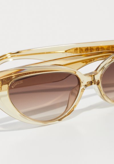 Gafas de sol doradas transparentes con forma de ojo de gato y lentes marrones degradados, con brazos elegantes y detalles del logo en la montura.