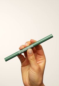 Crayon eyeliner vert tenu entre des doigts, présentant un design cylindrique lisse et un texte imprimé blanc sur le côté.