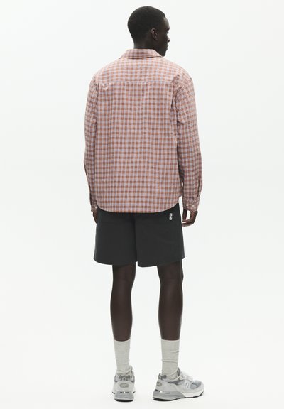 Homme vu de dos portant une chemise à manches longues à carreaux orange et blanche, un short noir, des chaussettes grises et des baskets blanches sur un fond uni.