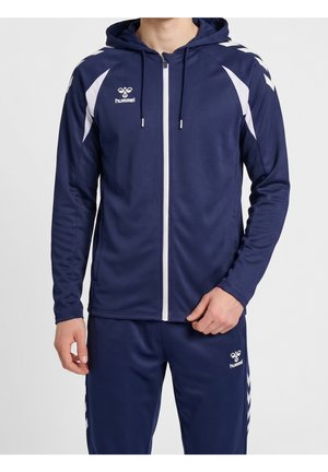 Mann trägt marineblauen Sportanzug mit weißen Chevron-Mustern, Kapuze und Frontreißverschluss, zeigt hummel-Logos auf Oberteil und Hose.