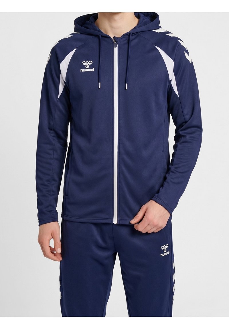 Homme portant un survêtement de sport bleu marine avec des motifs chevrons blancs, capuche et fermeture éclair à l'avant, montrant les logos hummel sur le haut et le pantalon.