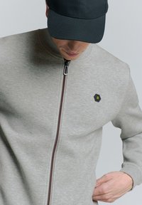 Lichtgrijze zip-up sweatshirt met een hoge kraag, voorzien van een hexagonaal logo en een rode accenten langs de rits. Soepele stoftextuur.