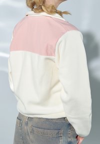 Henkilö, jolla on päällään vaaleanpunainen ja kermanvärinen fleecepaita sekä siniset farkut, selin päin, punotut vaaleat hiukset.