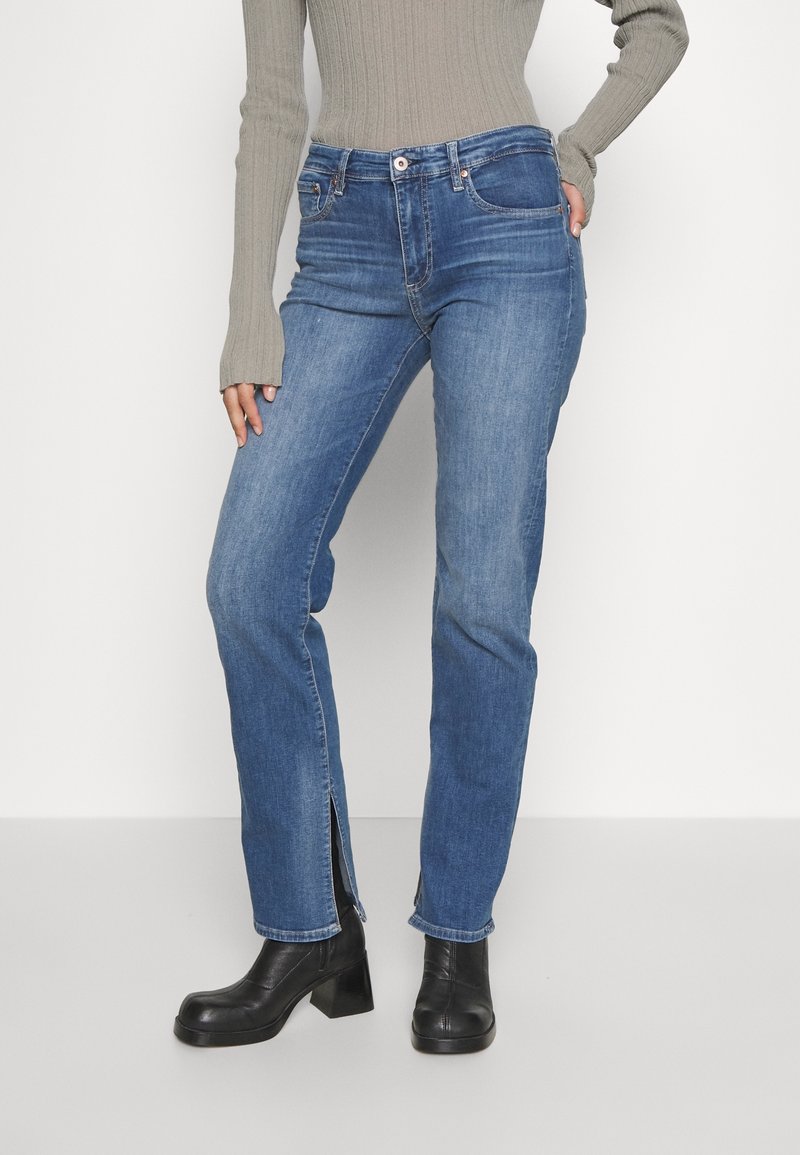 Personne portant un jean bleu droit avec des fentes à l'avant, associé à des bottes noires à talons épais et un haut gris côtelé à manches longues.