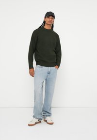 Donkergroene gebreide trui met een ronde hals, gecombineerd met lichtblauwe recht gesneden jeans en witte sneakers. Model draagt een zwarte pet.