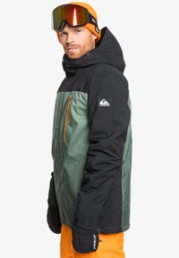 Chaqueta aislante verde y negra con un zipper y detalles en naranja. Cuenta con capucha, bolsillos y puños elásticos; la tela tiene una textura suave.