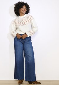 Balts džemperis ar rozā ģeometrisko rakstu uz piedurknēm, kombinēts ar platām zilo džinsu biksēm un brūnām potīšu zābakām. Redzami gludi tekstūri.