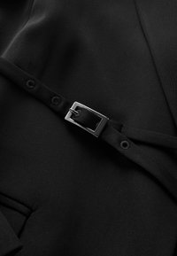 Czarna tkanina o gładkiej teksturze z zapięciem na klamrę i pasek, z dodatkowymi detalami w postaci guzików, podkreślającymi zorganizowany design.