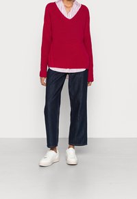 Rode gebreide trui met een V-hals, gelaagd over een wit en roze gestreept shirt, gecombineerd met een donkere wijde jeans en witte sneakers.