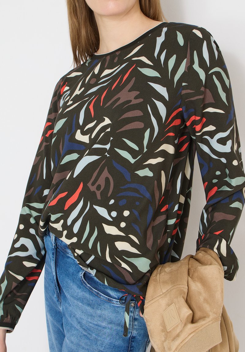 Femme portant un haut noir à manches longues avec des motifs abstraits rouges, bleus, verts et beiges, associée à un jean bleu, tenant une veste beige.