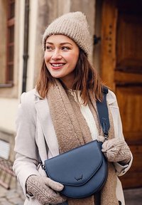 Modrá kožená crossbody taška s zakřiveným klopem a embosovaným logem, v kombinaci s béžovým pleteným šátkem a rukavicemi, na pozadí z dřeva.