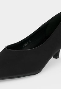 Chaussure femme noire à talon bas, bout fermé avec un avant-pointu et une finition lisse sur un fond clair.