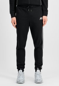 Zwarte trainingsbroek met witte zijstrepen, elastische tailleband met trekkoord, en een logo op de linker bovenbeen. Zachte stofstructuur.