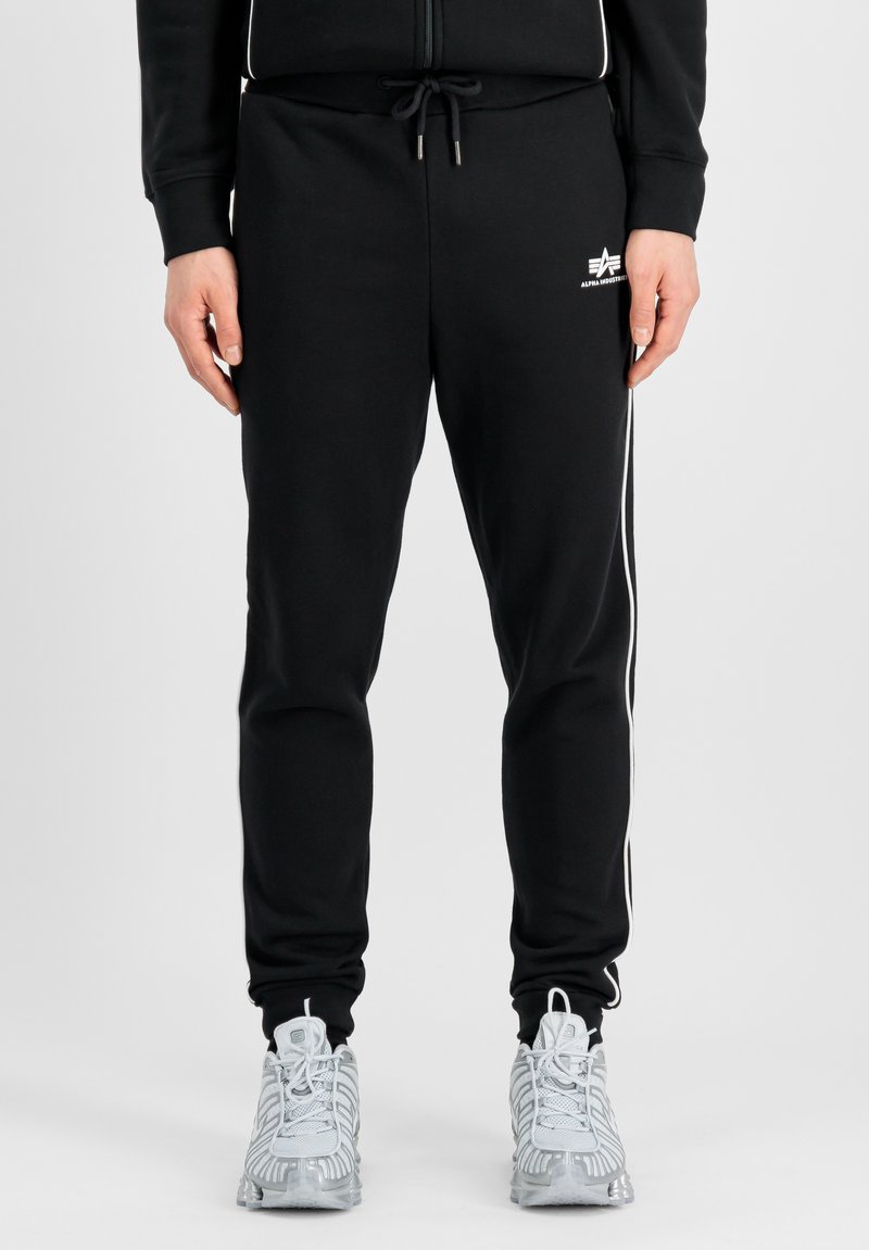 Zwarte trainingsbroek met witte zijstrepen, elastische tailleband met trekkoord, en een logo op de linker bovenbeen. Zachte stofstructuur.