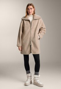 Beige fleecejas met een brede kraag en een ritssluiting aan de voorkant, gecombineerd met zwarte leggings en witte winterlaarzen met gestructureerde accenten.