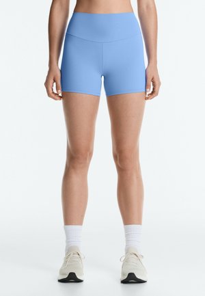 Femme portant un short de sport taille haute bleu clair, des chaussettes blanches et des chaussures de course couleur crème, debout avec les bras détendus le long du corps.