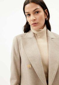 Beige ullblandad kavaj med slagkrage, dubbelknappar och skräddarsydd passform, över ett ljust beige polokrage-garn.