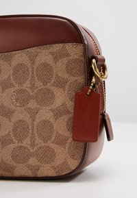 Ruskea nahkainen olkalaukku, jossa on tunnusomainen monogrammikuosi, kullanväriset metalliosat ja vastaava merkki, jossa on kohokuvioitu merkin nimi.