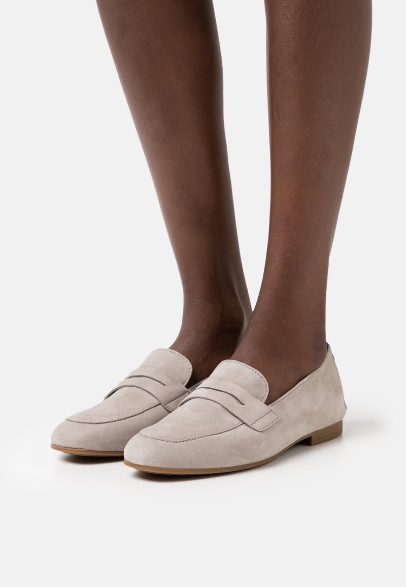 Mocassins en daim beige clair avec détail de lanière à penny, portés sur des jambes brun foncé, présentés sur un fond clair uni.