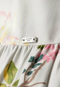 Detailní záběr na bílou látku s květinovým potiskem a malou kovovou značkou označenou "SUPER DRY", přišitou blízko švu.