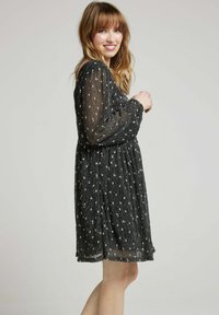 Robe noire avec de longues manches transparentes, présentant un motif texturé et de petits détails floraux blancs. Silhouette évasée qui tombe au-dessus du genou.