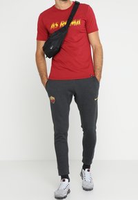 Camiseta deportiva roja con el logotipo de AS Roma en amarillo, combinada con pantalones de chándal gris oscuro que presentan un logotipo y acentos amarillos. Mochila cruzada negra y zapatillas.