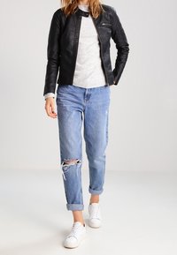 Svart läderjacka med texturerade axlar, grå tröja under, slitna ljusblå jeans och vita sneakers, stående på en neutral bakgrund.