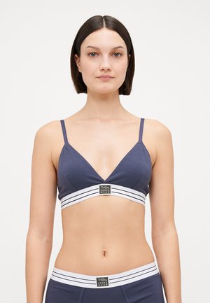 Navy fabric bralette with a deep V neckline, thin adjustable straps, and a wide white striped band featuring a logo at the center.
