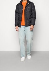 Czarna kurtka puchowa z wypukłym napisem, zapinana na zamek z przodu, z kołnierzem na zatrzaski, noszona na jasnopomarańczowej koszuli, jasnoniebieskich dżinsach i białych butach.