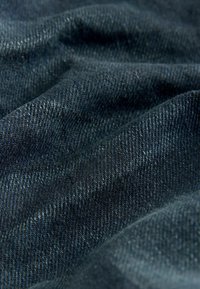 Țesătură de denim albastru închis, cu țesătură twill vizibilă, pliată și texturată, evidențiind variații subtile de nuanță și un luciu ușor.