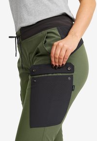 Woman wearing olive green pants with a large black side pocket, hand lifting the pocket flap with dark nail polish visible.