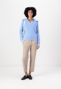 Pulover tricotat bleu deschis cu guler larg, asortat cu pantaloni beji și mocasini negri. Prezintă un mic logo pe pulover.
