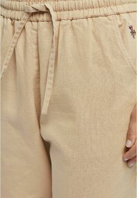 Beige bomullsjoggers med elastisk midja, snörning och en liten broderad detalj. Slät textur med avslappnad passform.