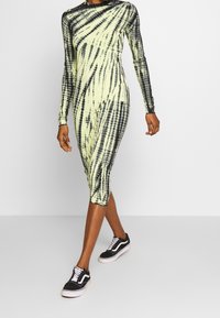 Šaty s dlouhým rukávem v žluté a černé barvě s tie-dye vzorem. Přiléhavý střih s vysokým výstřihem. Kombinováno s černými teniskami. Hladký povrch.