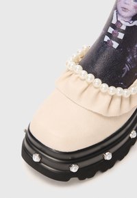 Chunky beige shoe with black sole decorated with pearl studs, ruffled leather trim, and a printed sock featuring a person in striped clothing.
