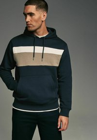 Marine sweatshirt med hætte, der har tre vandrette striber: beige og hvidt henover brystet. Inkluderer frontlomme og ribbede ærmer.