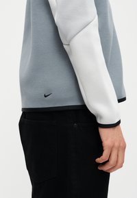 Seitenansicht einer Person, die ein graues und weißes Nike-Langarmshirt mit schwarzen Hosen trägt, wobei der Nike-Swoosh im Hüftbereich sichtbar ist.