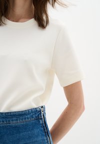 Tricou crem cu mânecă scurtă dintr-un material moale, cu decolteu rotund. Potrivit cu o fustă din denim cu talie înaltă, care are un fermoar vizibil.