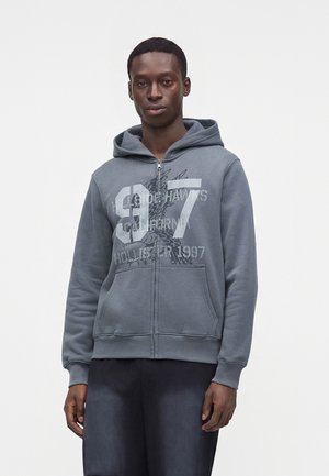 Jeune homme portant un sweat à capuche bleu zippé avec un grand 97 et un motif d'aigle, debout avec une posture détendue sur un fond blanc uni.