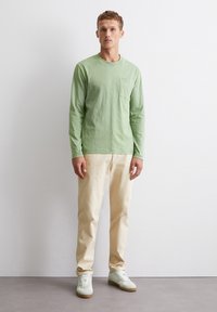 Langärmliges grünes Baumwoll-T-Shirt mit einer Brusttasche, kombiniert mit beigen Hosen und hellgrünen Turnschuhen. Lässige, lockere Passform.