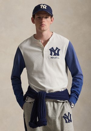 Νεαρός άνδρας που φοράει λευκό και μπλε μακρυμάνικο μπλουζάκι New York Yankees, καπέλο που ταιριάζει, γκρι φόρμες και μπλε πουλόβερ δεμένο γύρω από τη μέση του.