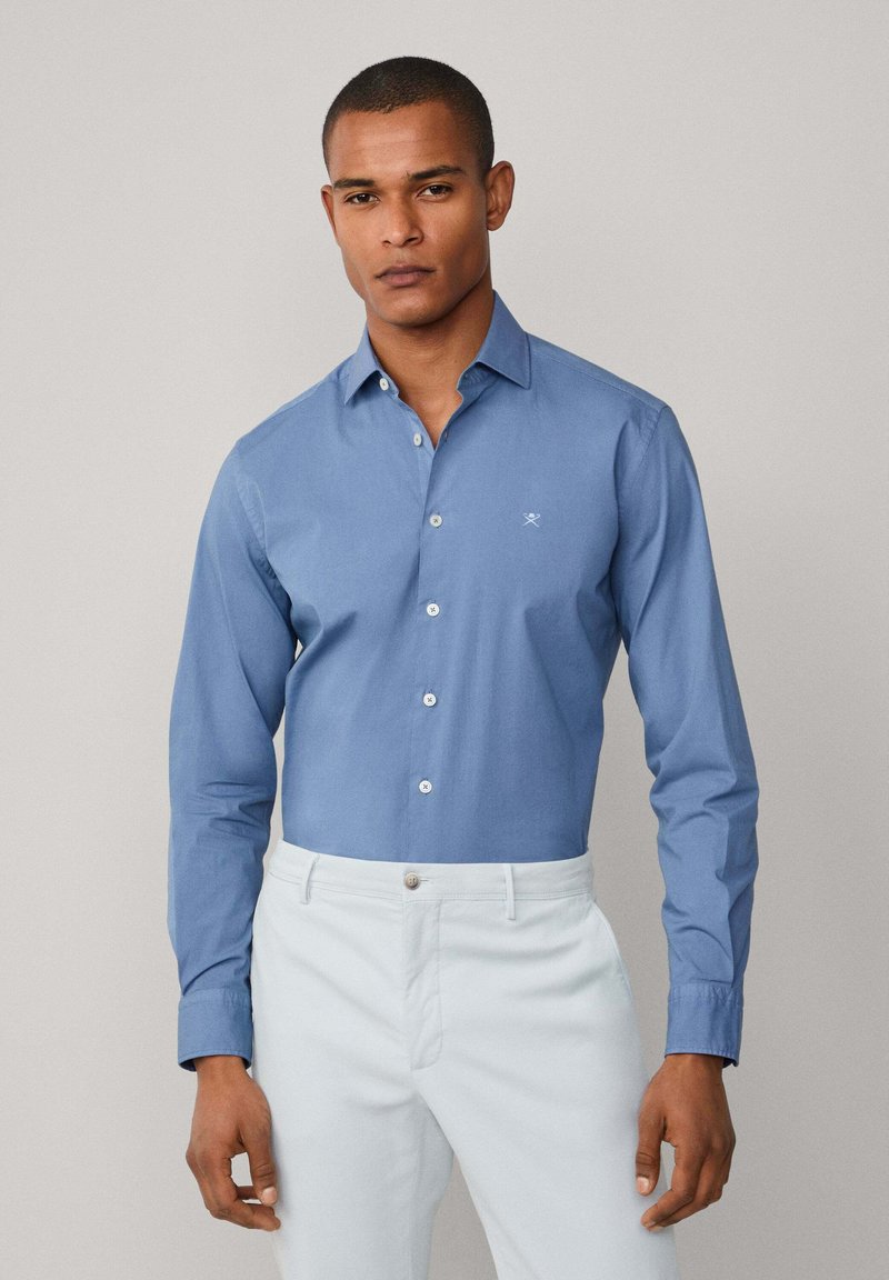 Camisa de botones azul claro con mangas largas y collar clásico. Presenta un sutil logo bordado en el pecho. Material de algodón.