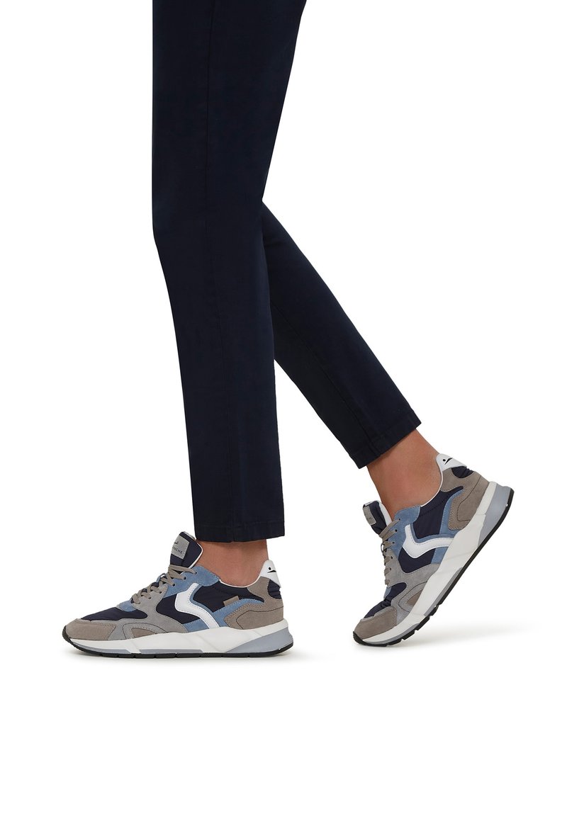 Grå och blå sportskor med en kombination av mocka och nätmaterial, vita detaljer och en strukturerad sula, bärs med mörka, korta byxor.